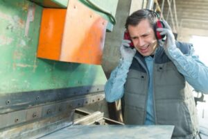 Factory worker suffering hearing loss from loud machinery noise in industrial workplace.