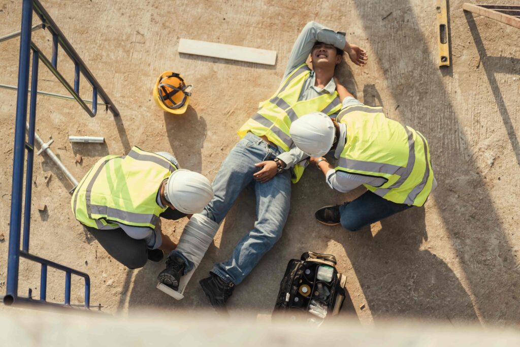 Construction worker injured on job site receiving first aid from coworkers in safety vests and helmets